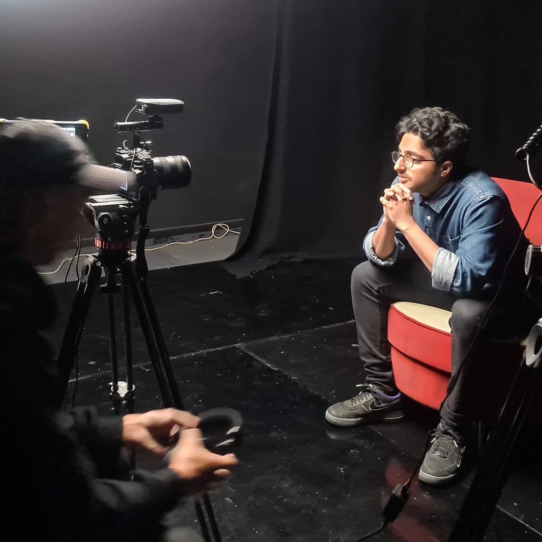 Harsha sits in a red chair and leans forward, he's being filmed in a black room with a guy in a black baseball cap